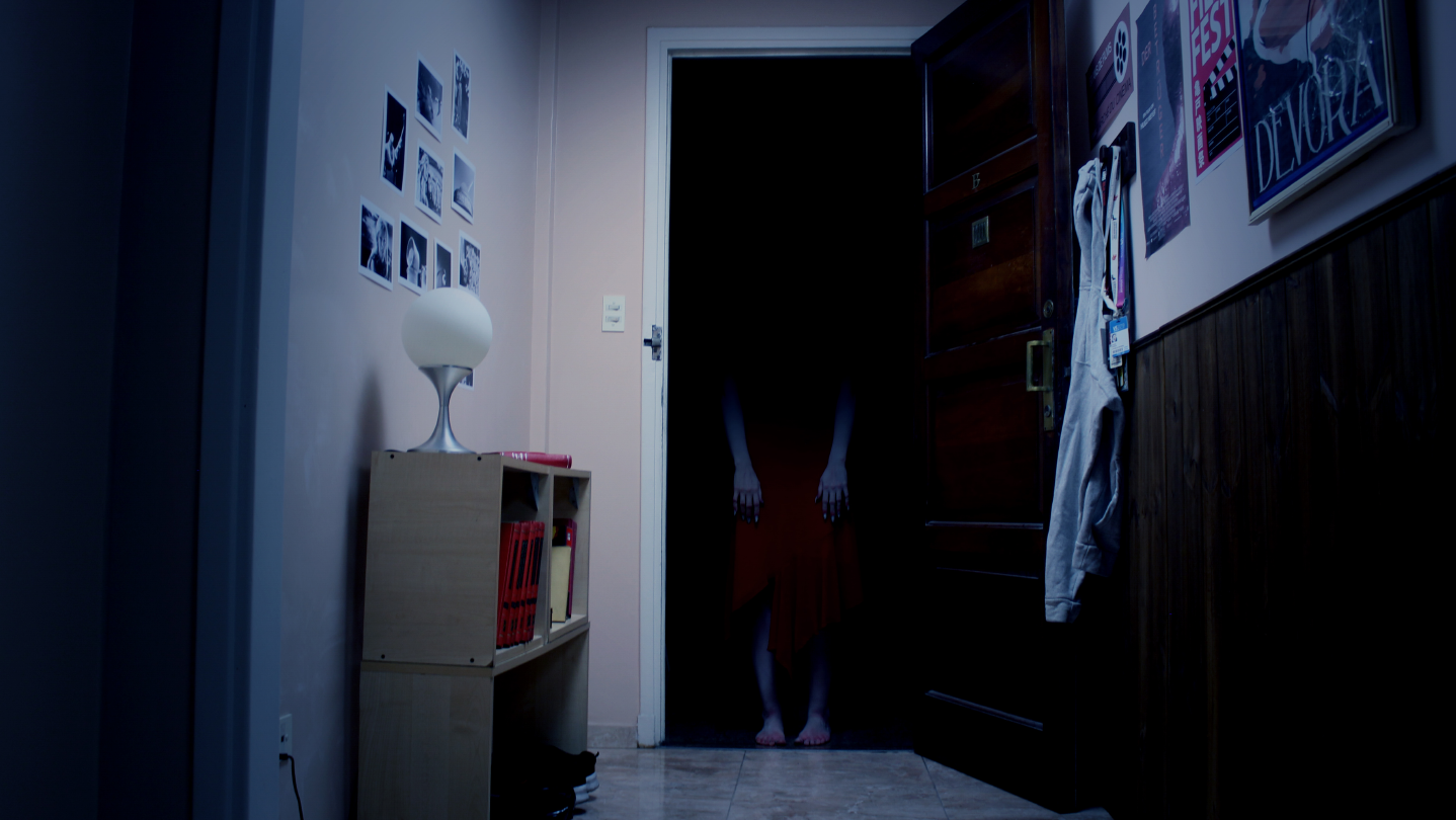 Tenebris Somnia key art: a cold blue hallway where a figure in a red dress stands in a black doorway, with film stills pinned to the wall and a white lamp on a shelf of red books on the left, and an open wooden door beside DEVORA film posters and a hanging white coat on the right.